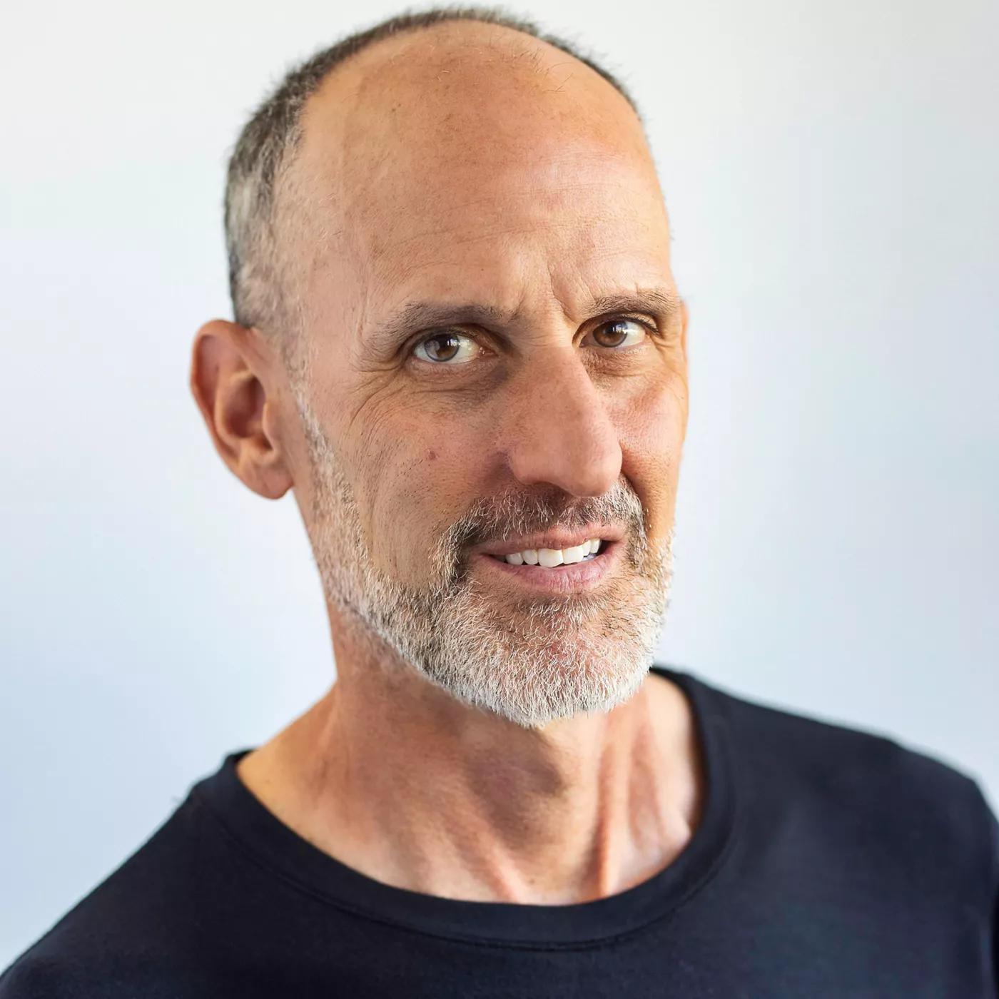 A middle-aged man wearing a plain black shirt is photographed against a neutral light background. The image focuses on the upper torso and shoulders, showcasing a casual and minimal style. The setting is simple and clean, emphasizing the subject's attire and posture.